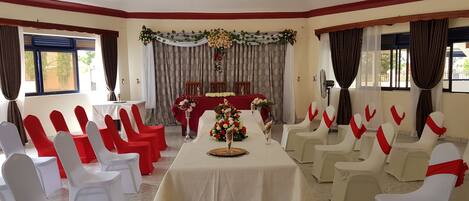 A banquet hall with rows of white and red chairs, a long table with floral centerpieces, and a backdrop with curtains and floral decorations.