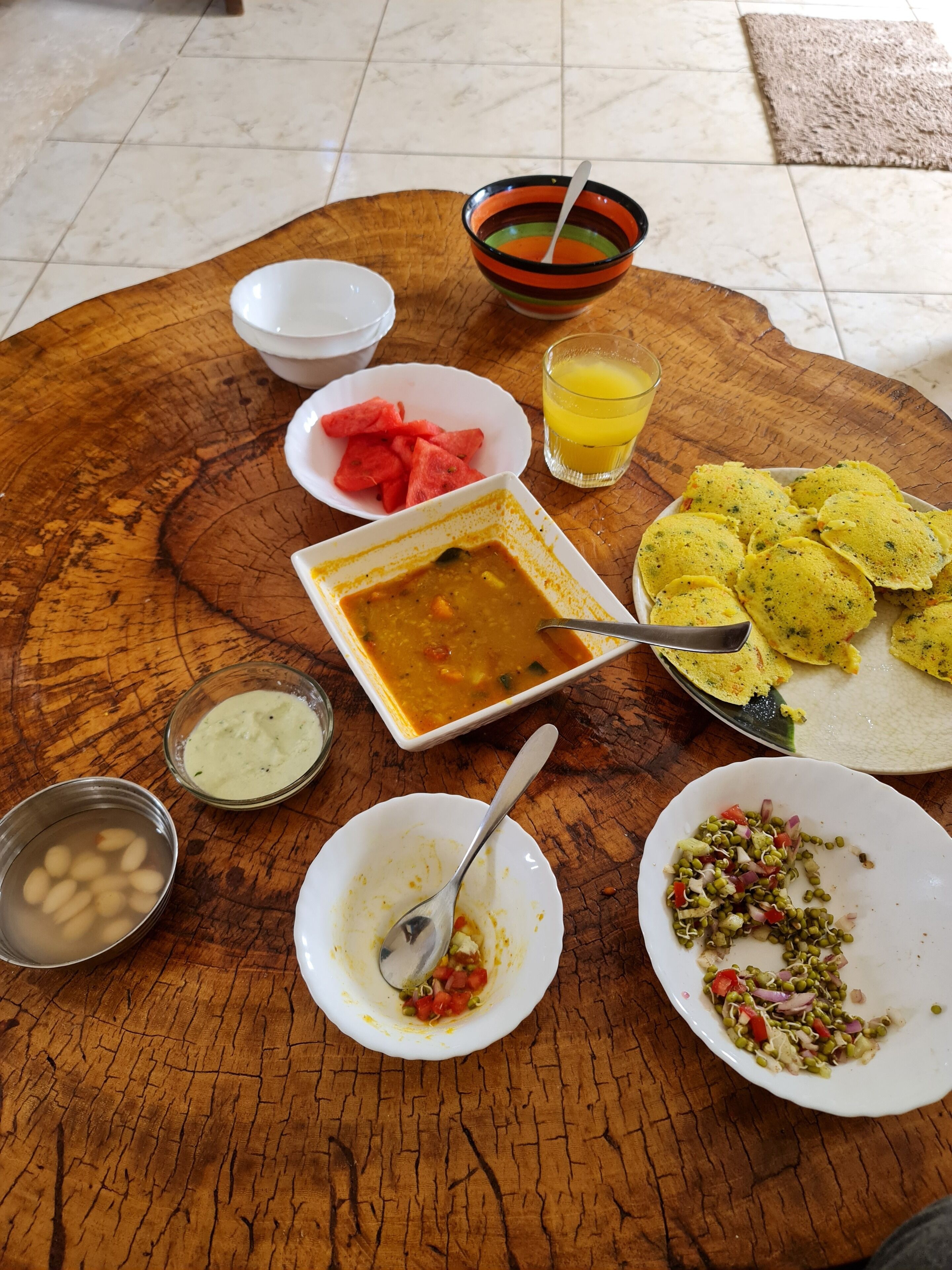 Une table en bois sur laquelle on trouve différents plats, notamment un bol de soupe, des tranches de pastèque, un verre de jus d’orange et une assiette de galettes.