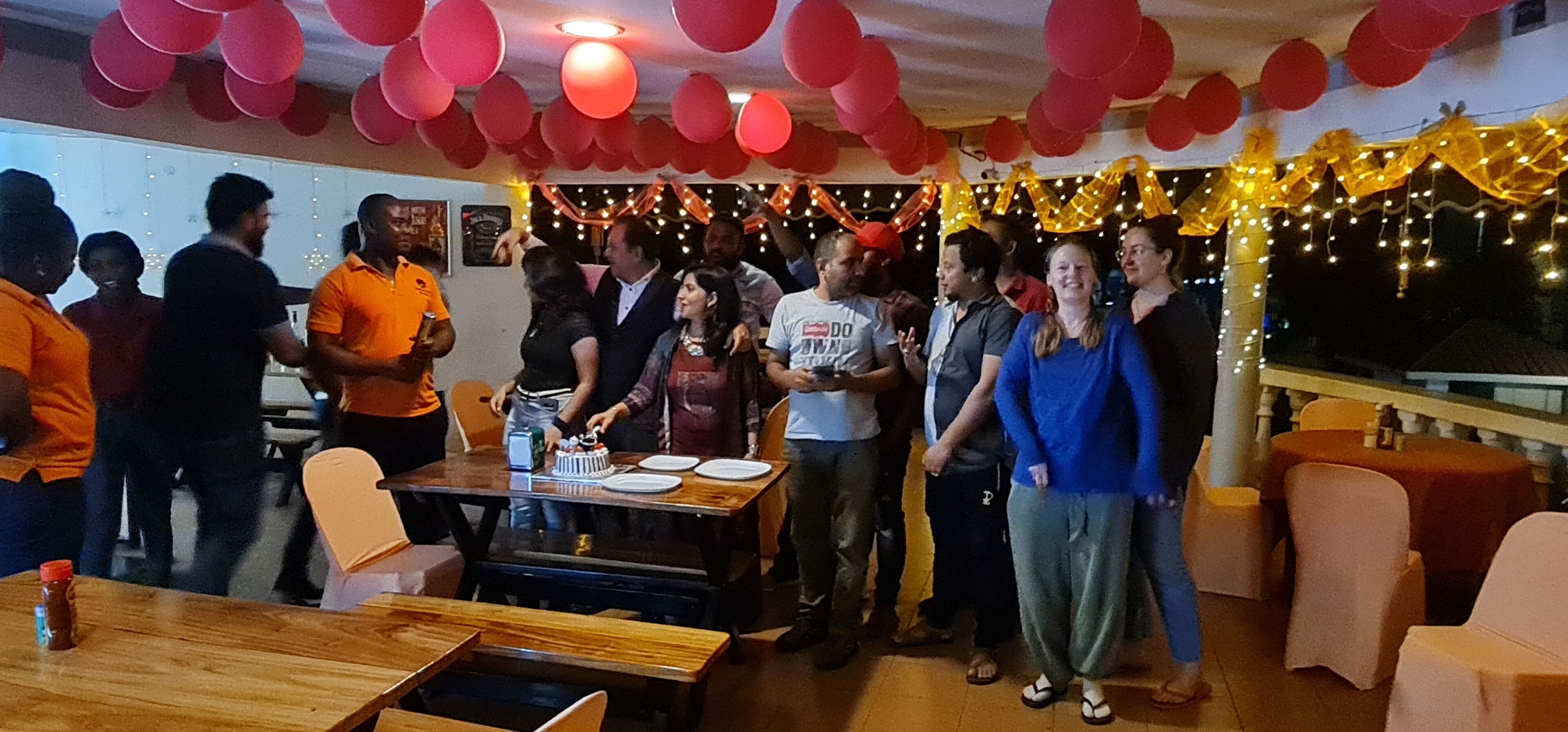 A group of people gathered around a table with a cake, in a room decorated with red balloons and string lights.