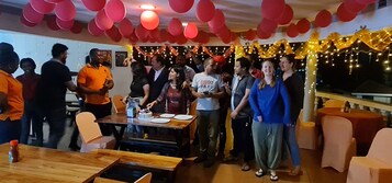 A group of people gathered around a table with a cake, in a room decorated with red balloons and string lights.