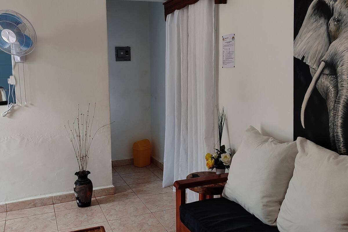 A room with a bed, a standing fan, a small table with a vase of flowers, and a picture of an elephant on the wall.