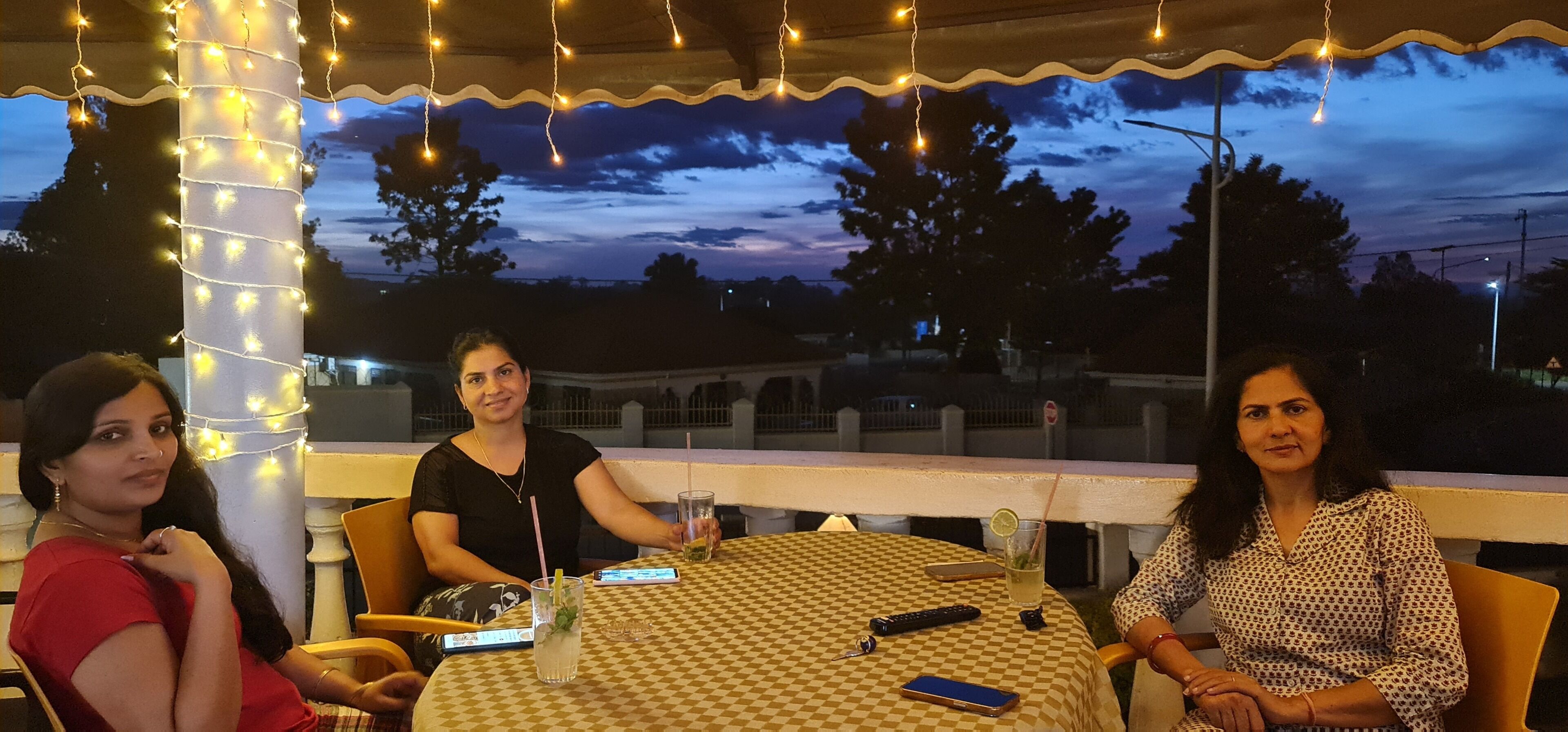 A group of people seated at an outdoor table with a checkered tablecloth, under a canopy with string lights, with a view of trees and a building in the background.