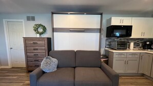 Un salon moderne avec un canapé gris, un meuble en bois et un coin cuisine équipé d’appareils électroménagers.
