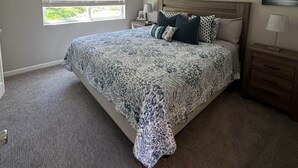 Un dormitorio con una cama grande, un ventilador de techo, dos ventanas con persianas, una mesita de noche y un cuadro en la pared.