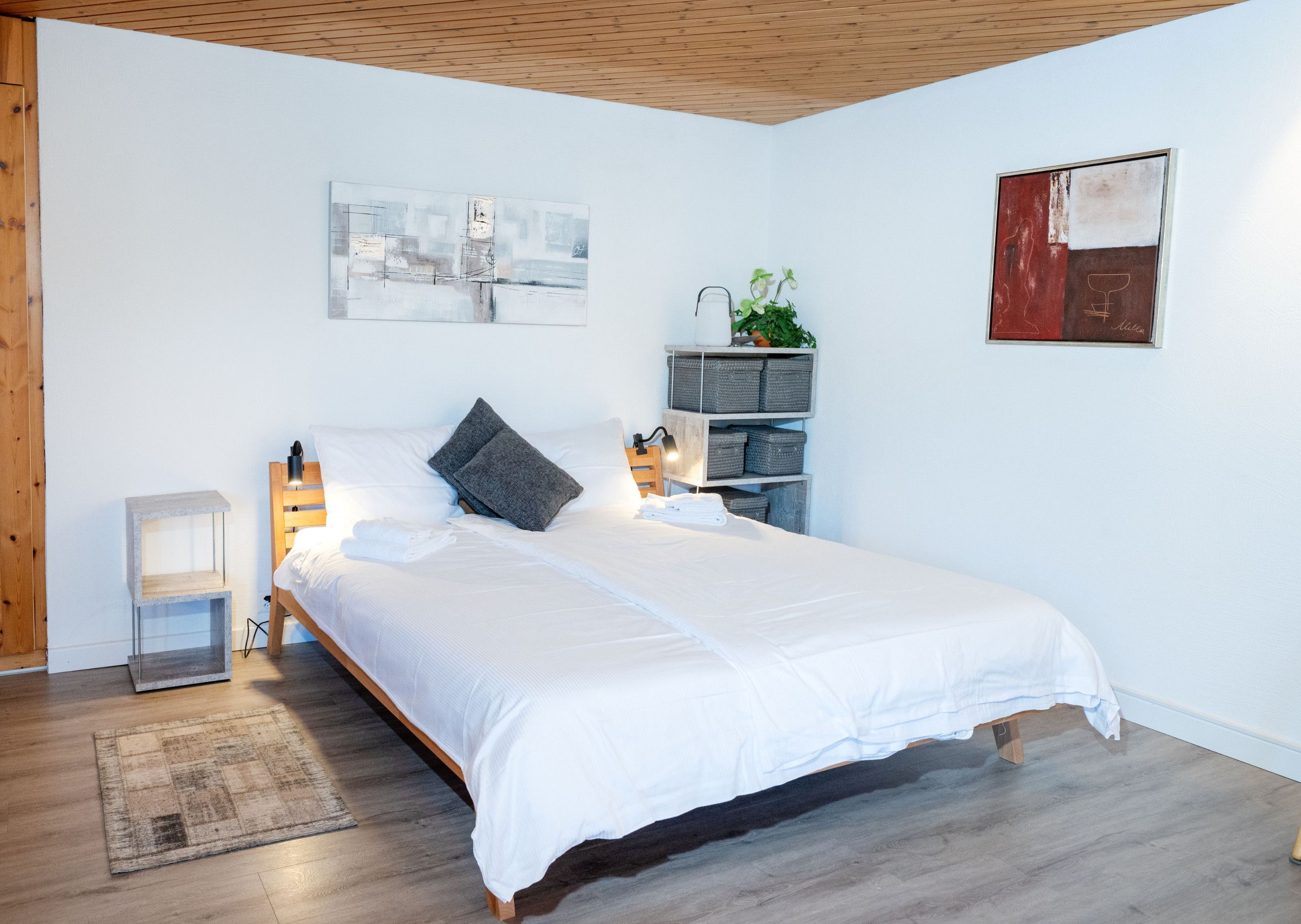 Un dormitorio con cama, mesitas de noche, una estantería de pared con plantas y obras de arte.