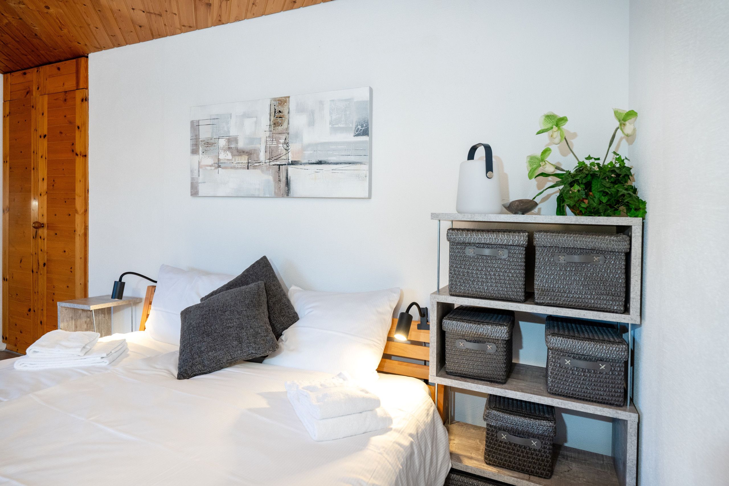 A bedroom with a bed, pillows, a bedside table, a lamp, a wall art piece, a plant, and storage baskets.