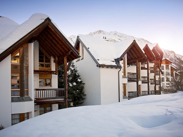 Un bâtiment enneigé, doté de balcons en bois, avec une chaîne de montagnes en arrière-plan.