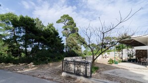 Un carril pavimentat que condueix a una porta de pedra, envoltat de vegetació densa i un edifici amb una zona de terrassa coberta.