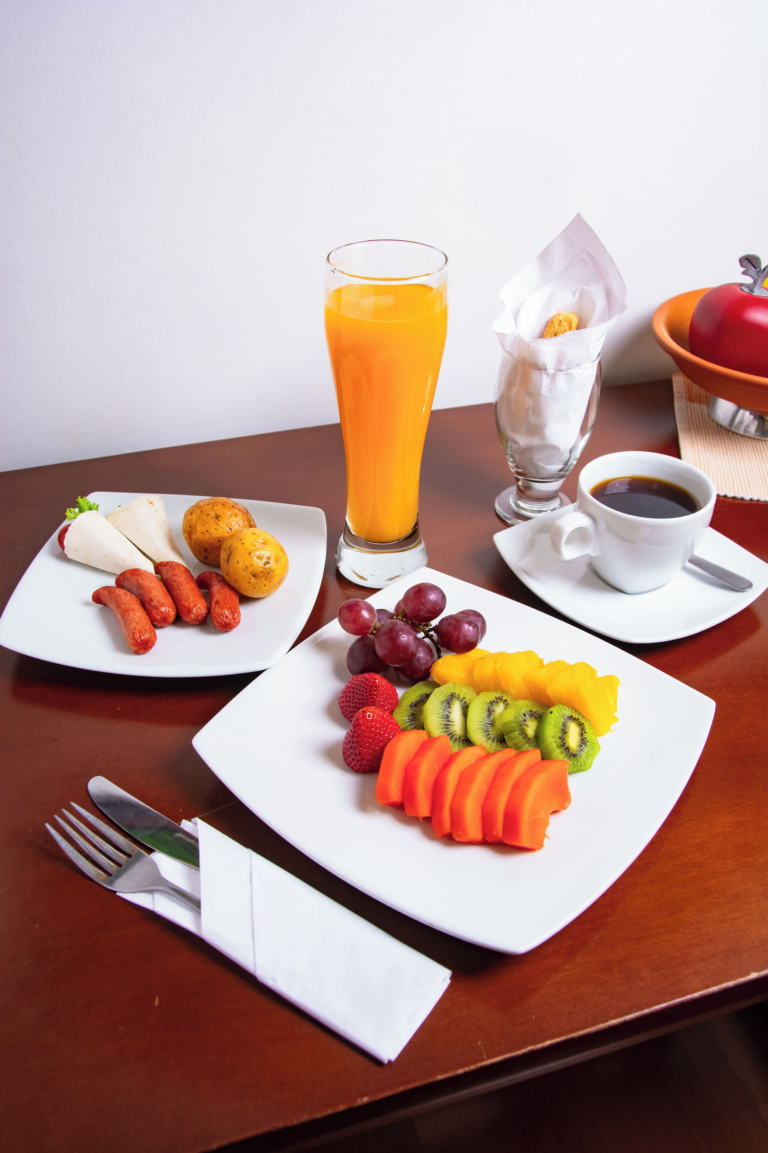 A breakfast setting with plates of fruit, sausages, and a glass of orange juice.