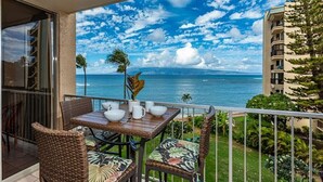 A balcony with wicker furniture, a table with white cups and a plant, overlooking a coastal view with buildings and greenery. -   Dlx OCEANVIEW Maui  Studio..Updated!
West Maui. AC. Royal Kahana. (Lahaina)