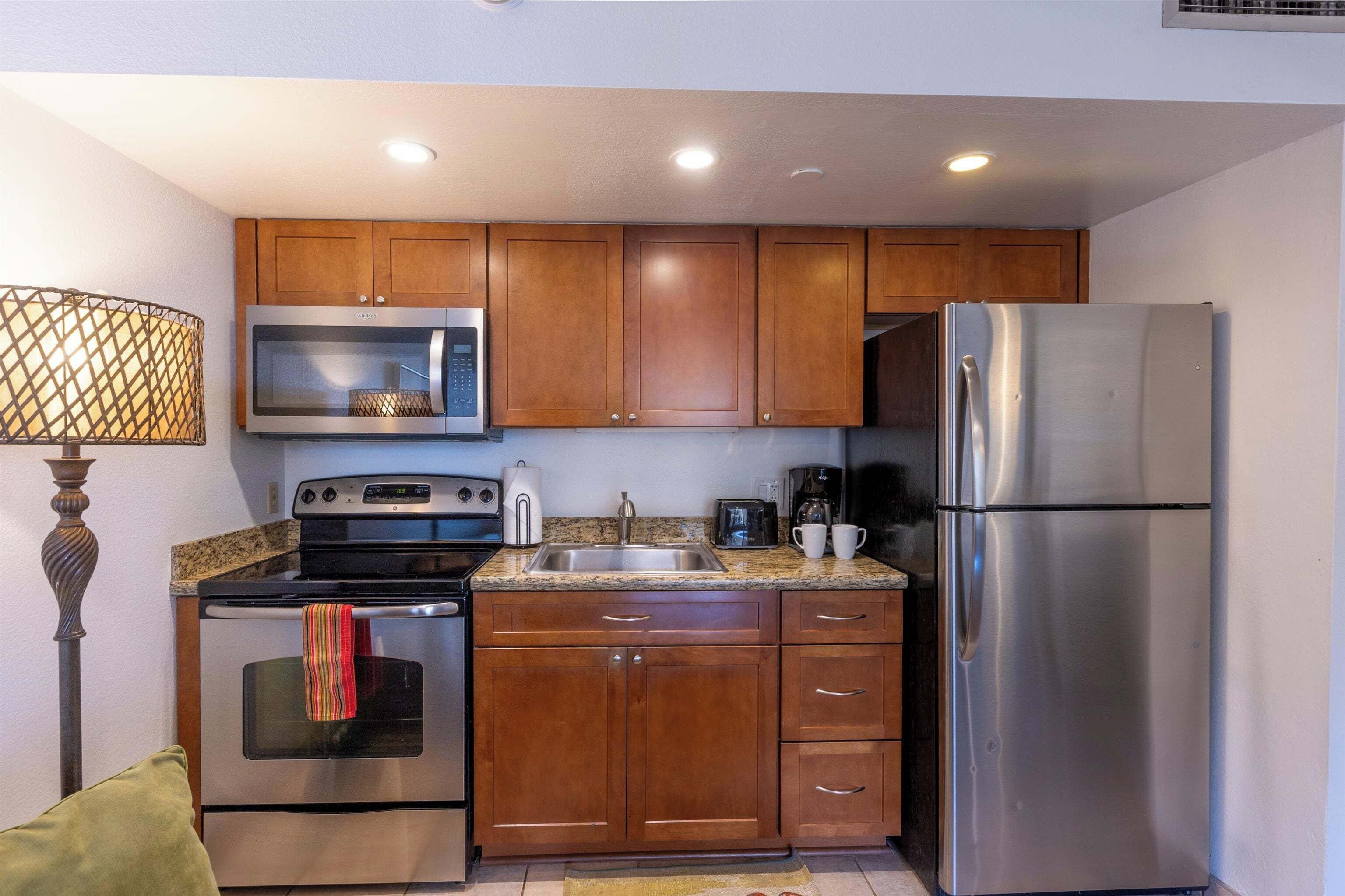 A compact kitchen with wooden cabinets, stainless steel appliances including a microwave, oven, and refrigerator, and a granite countertop.