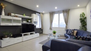 Een moderne woonkamer met een flatscreen televisie, witte kasten, een blauwe bank en een groot raam met gordijnen.