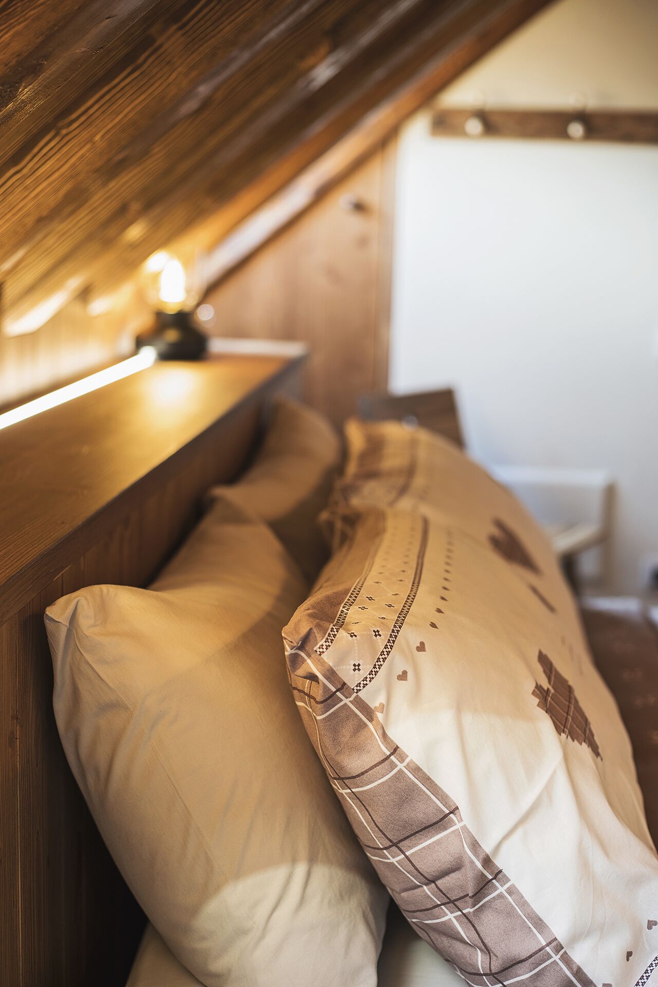 A cozy bedroom with a wooden headboard, a bedside table with a lamp, and two pillows with a checkered pattern.