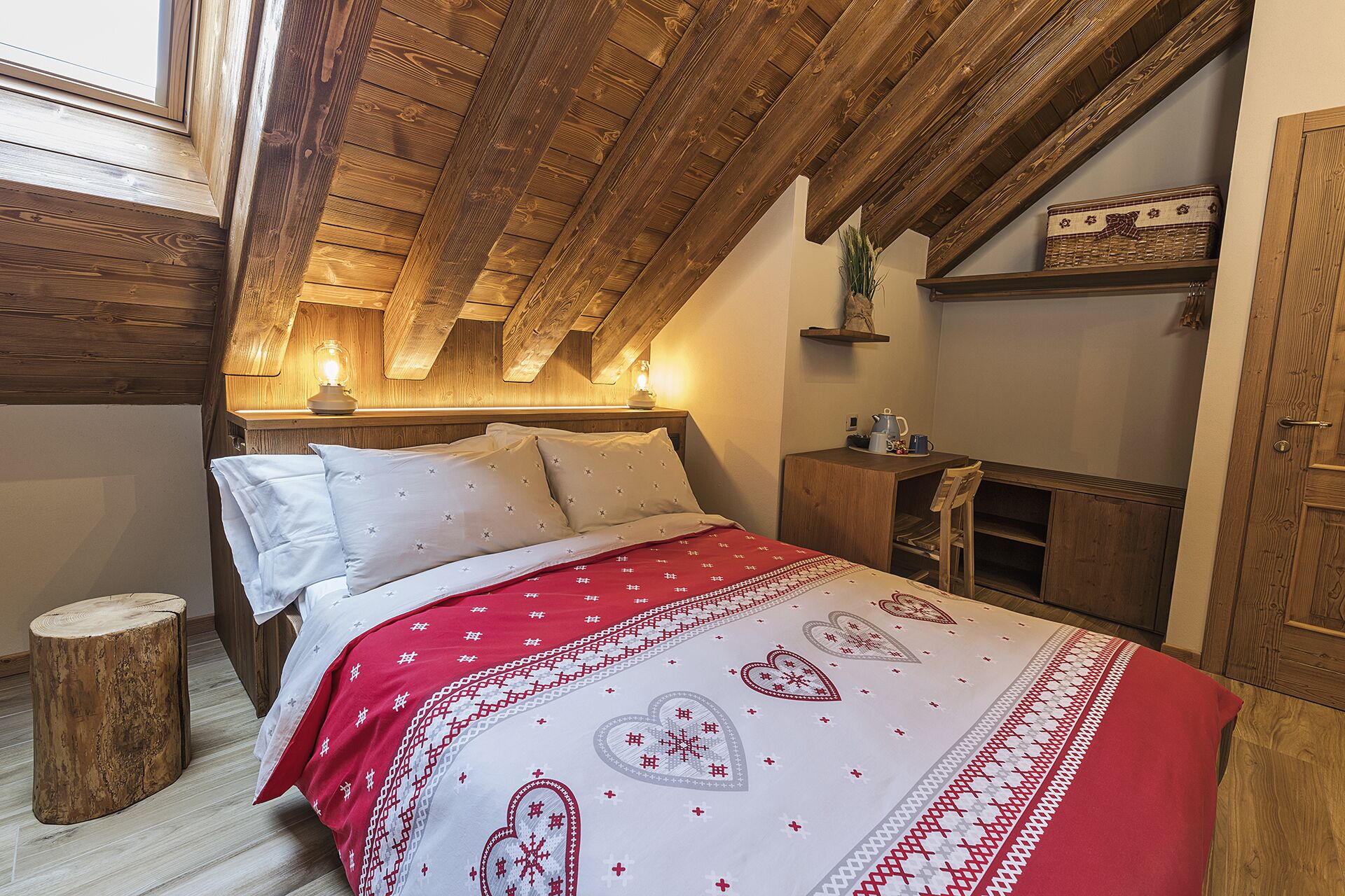 A cozy bedroom with a sloped ceiling, wooden beams, a bed with a red and white patterned bedspread, and a small wooden stool.