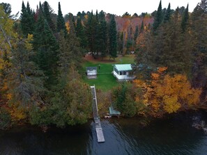 Una cabaña con un muelle que se adentra en un lago, rodeada de un bosque denso con follaje otoñal.