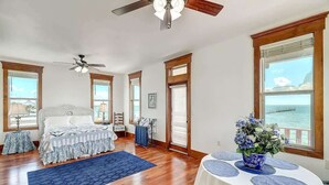 Un dormitorio con cama, ventilador de techo, ventana grande con vistas y una mesa con un jarrón de flores.
