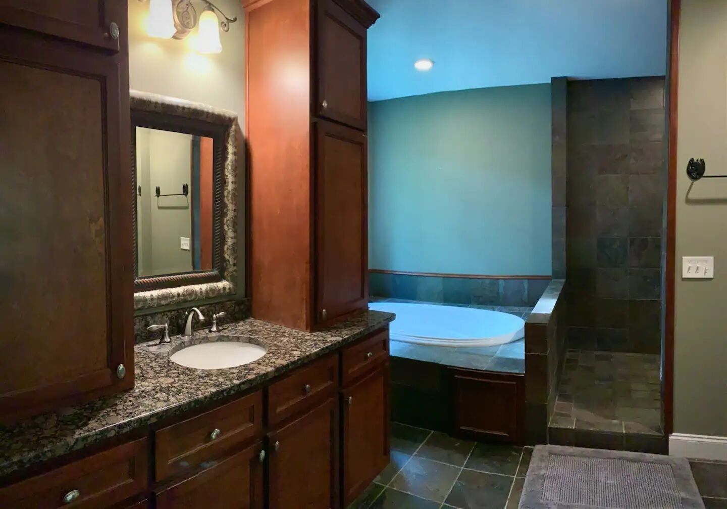 Une salle de bain dotée d’un lavabo avec un plan de travail en granit, de meubles en bois, d’un grand miroir et d’un espace douche aux carreaux en pierre.
