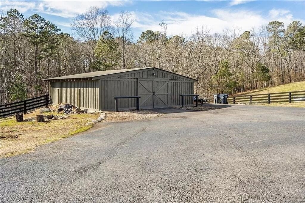 A large, single-story building with a flat roof, likely a barn or storage facility, situated on a paved driveway with a wooden fence on one side and a grassy area on the other.