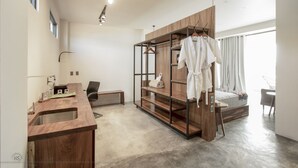 Een moderne hotelkamer met een houten opbergmeubel, een bed met houten hoofdbord, een kledingkast met daarin hangende kleding, en een groot raam.