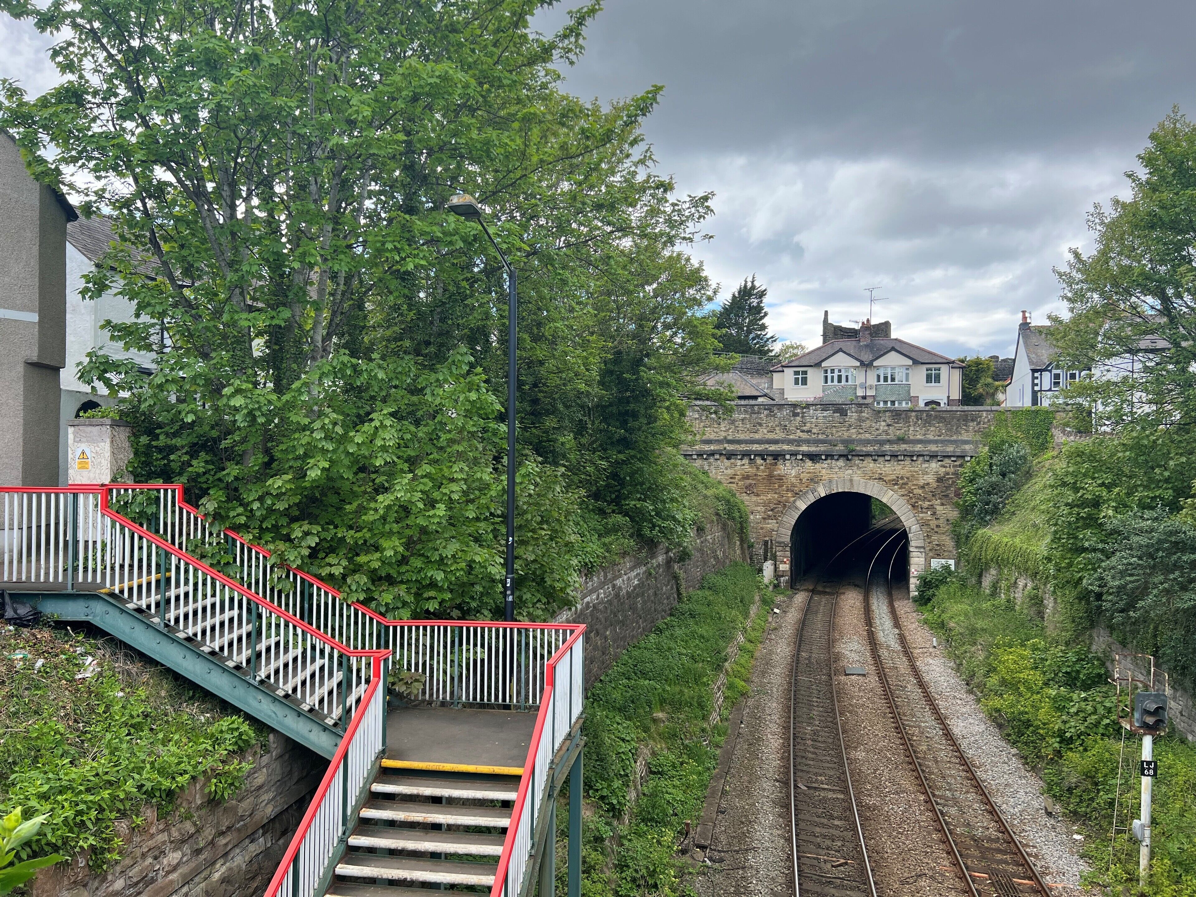 A railway tunnel entrance with a staircase leading down to the tracks, surrounded by greenery and residential buildings.
