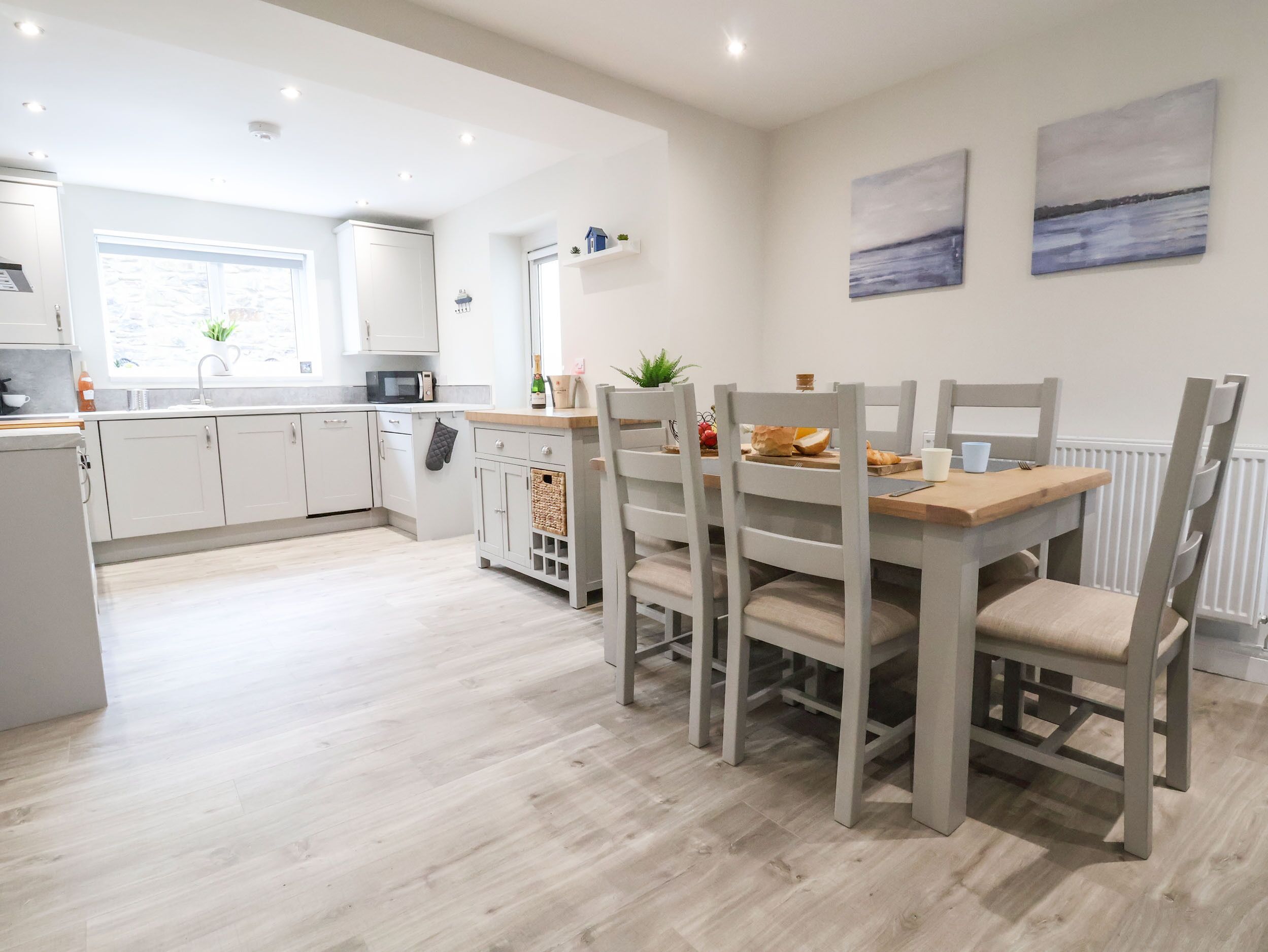 A modern kitchen with white cabinets, a dining table with chairs, and a view of the sea through the window.