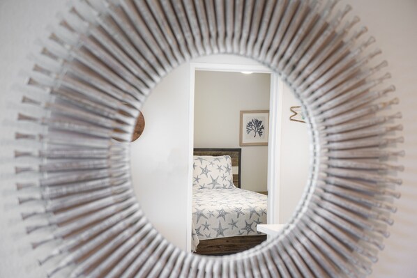 Un miroir rond au motif rayonnant, qui reflète une chambre à coucher avec un lit et un tableau encadré accroché au mur.