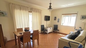 A living room with a sofa, a dining table with chairs, a TV, and a window with curtains. - Algarve, T2 apartment w/balcony, Armação de Pera (Armação de Pêra)