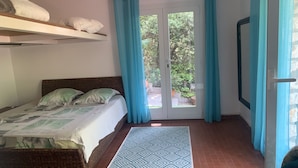 Un dormitorio con cama, ventana con cortinas, puerta y vista a follaje exterior.