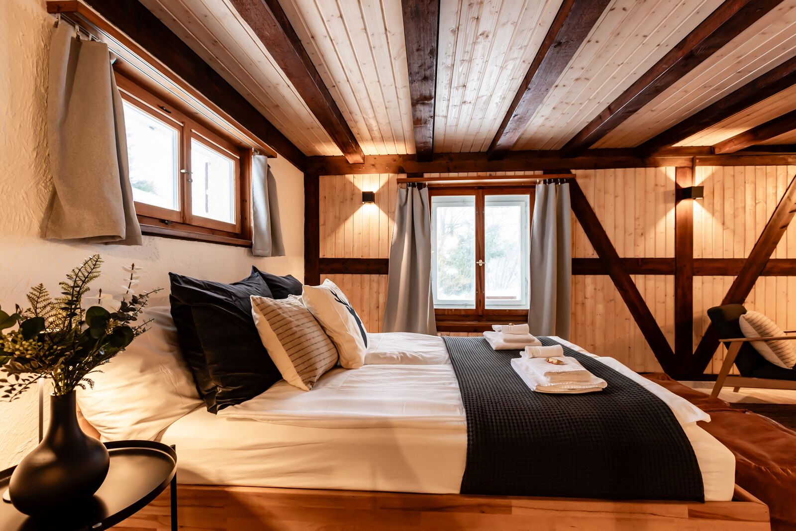 A cozy bedroom with a wooden ceiling, a large bed with multiple pillows, a bedside table with a vase, and a chair.