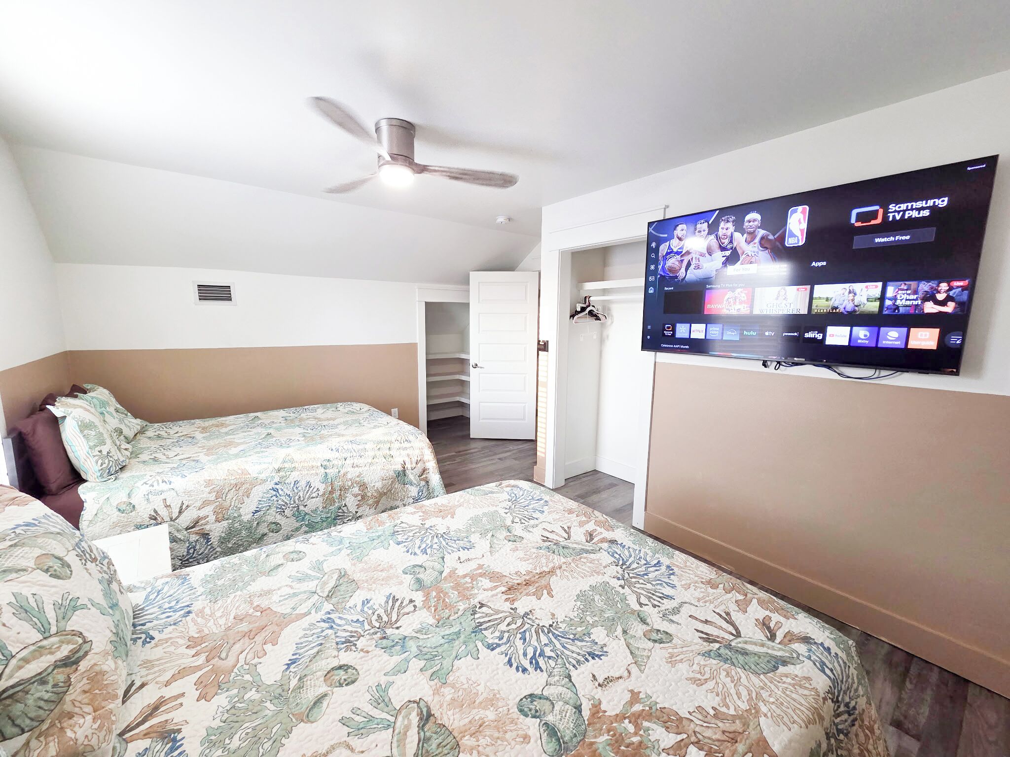 Habitación de hotel con dos camas, ventilador de techo y televisor montado en la pared mostrando diferentes canales.