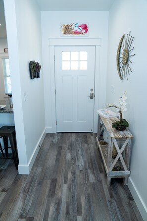 A hallway with a white door, a wooden bench with a vase of flowers, and a wall-mounted decorative piece.