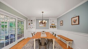 A dining room with a wooden table, chairs, and benches. There are built-in shelves with decorative items and books. A chandelier hangs from the ceiling, and the room has large windows with a view of the outdoors. - Berkshires Colonial on 2 Acres by Evergreen Home (Pittsfield)
