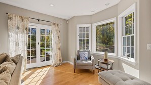 A living room with a sofa, a chair, and a bench. There are large windows with curtains, a wooden floor, and a ceiling light. - Berkshires Colonial on 2 Acres by Evergreen Home (Pittsfield)