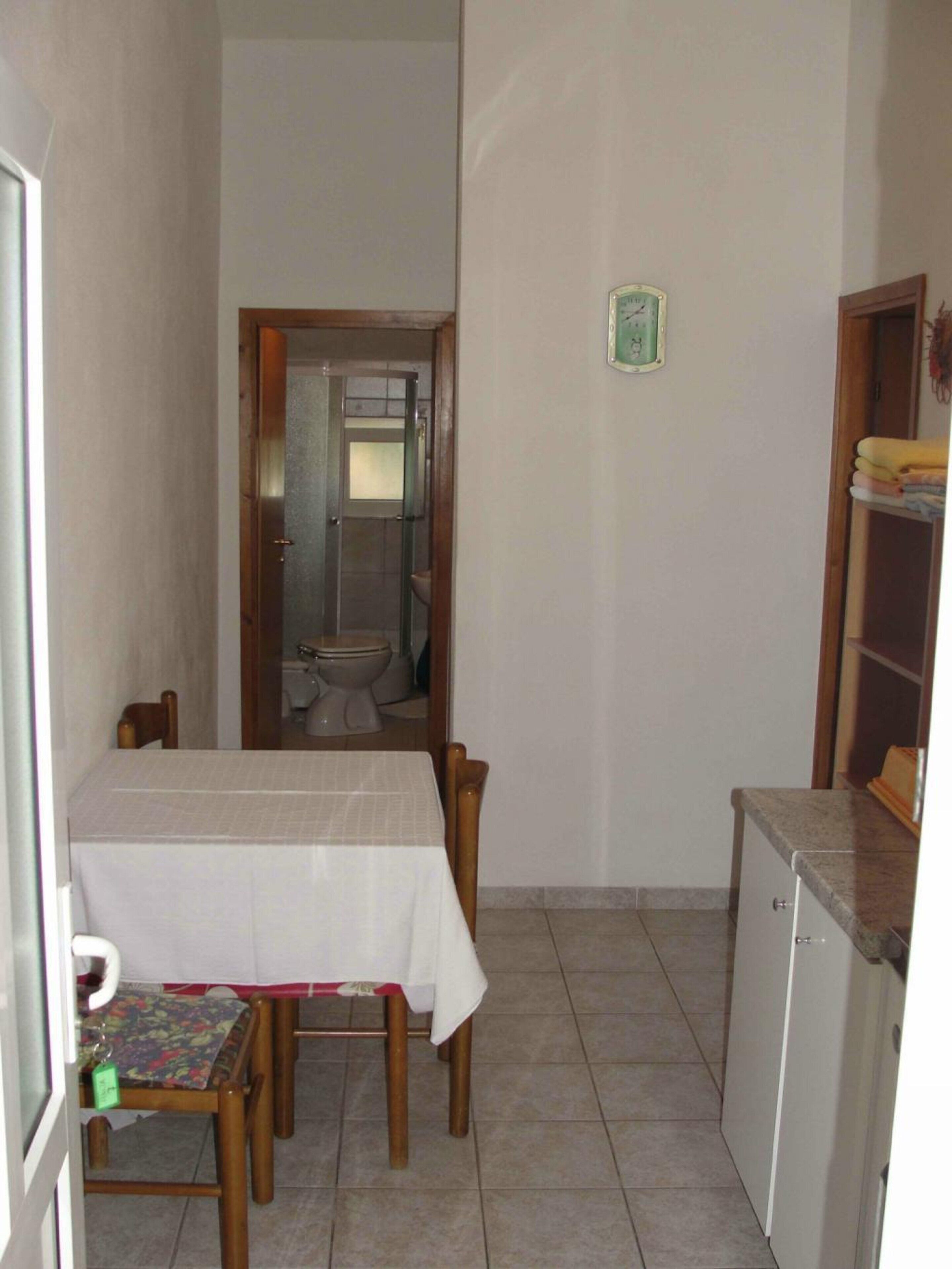 A small, narrow room with a dining table and chairs, a small refrigerator, and a toilet visible through an open door.
