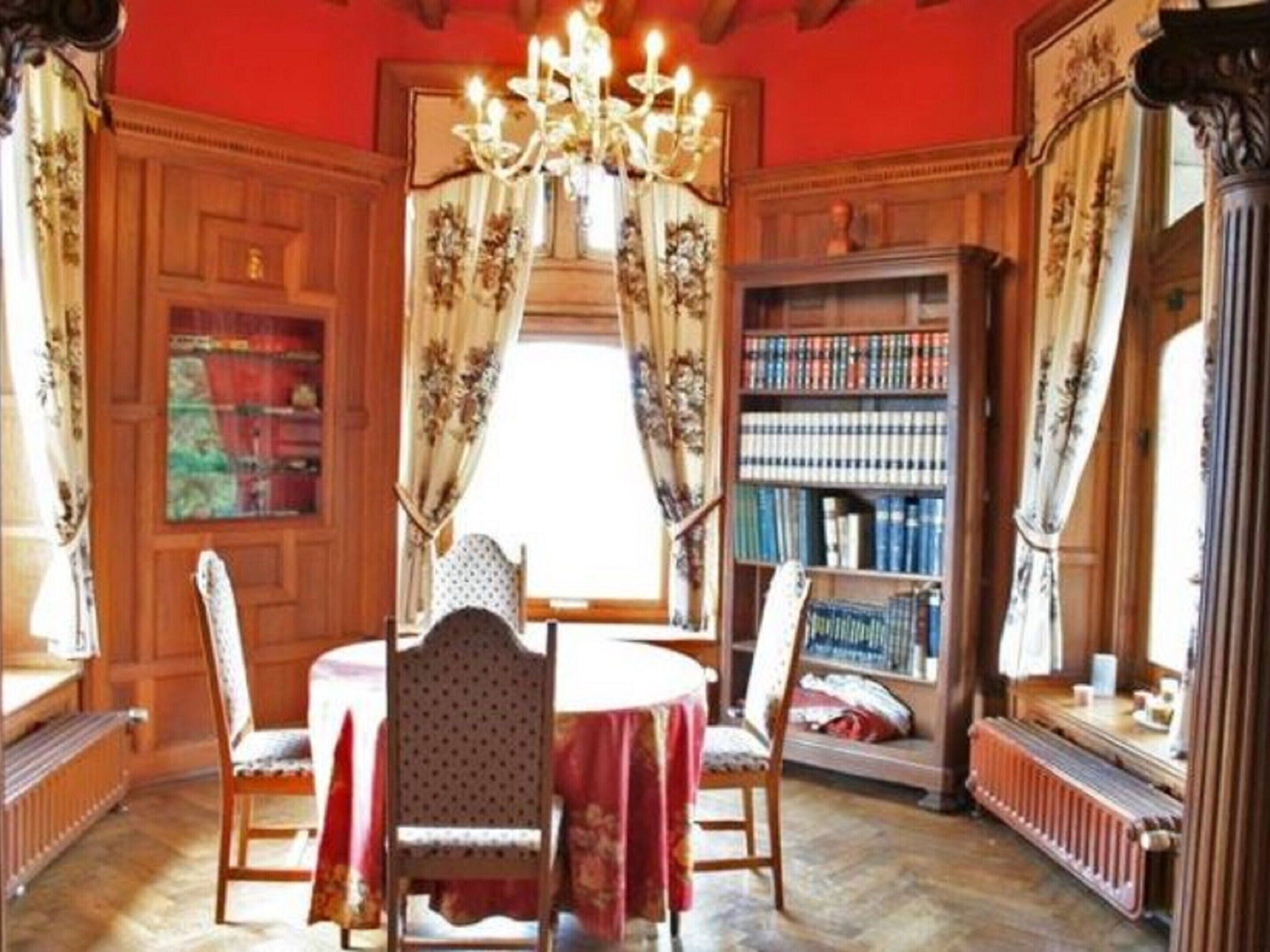 A room with a wooden paneled wall, a large bookcase filled with books, a chandelier, and a table with chairs.