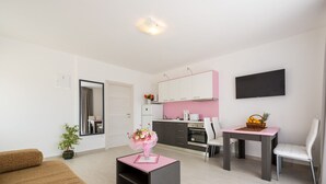 Ein modernes Wohnzimmer mit einem braunen Sofa, einem kleinen schwarzen Couchtisch mit pinkfarbener Tischplatte und einem weißen Sessel. Im Hintergrund ist eine Küche mit pinkfarbenen Schränken und weißen Geräten erkennbar.