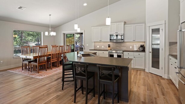 A modern kitchen with white cabinets, a large island, and a dining area with wooden chairs. - Manzanita - Hot Tub, Canal Front, Amazing Location! (Bend)