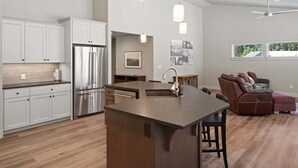 A modern kitchen with white cabinets, a stainless steel refrigerator, a central island with a sink, and a dining area with a brown leather couch. - Manzanita - Hot Tub, Canal Front, Amazing Location! (Bend)