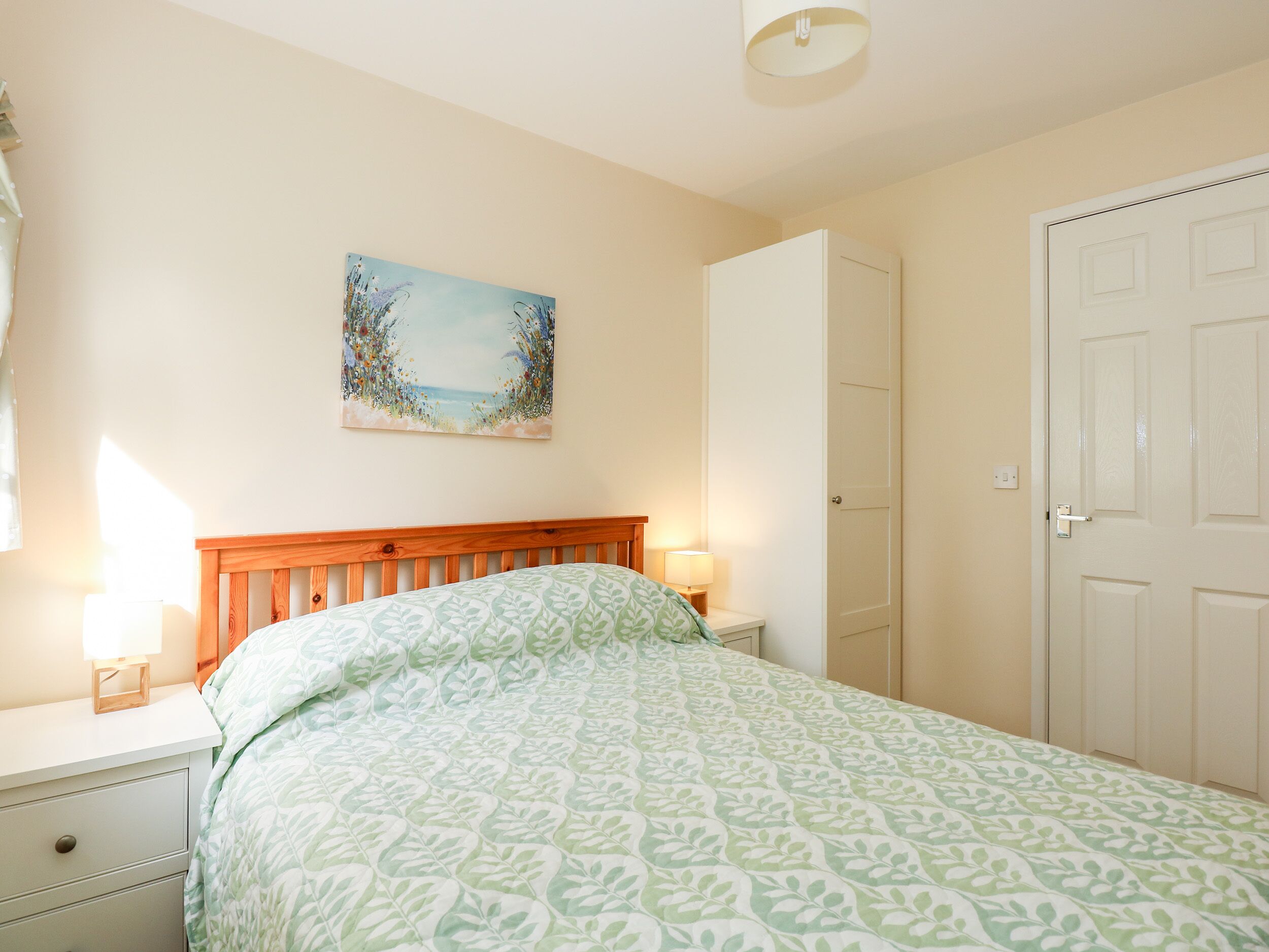 A bedroom with a wooden headboard, a bed with a green patterned bedspread, a nightstand with a lamp, and a door to the right.