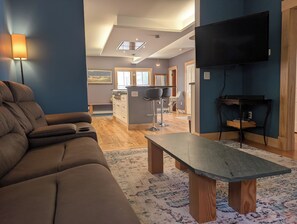 A modern living room with a sofa, a dining area with chairs, a kitchen with a bar, and a TV mounted on the wall.