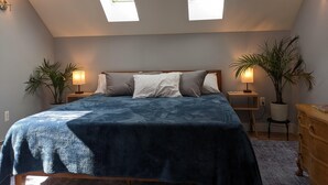 Un dormitorio con tragaluz, una cama con manta azul, dos mesitas de noche con lámparas y un aparador de madera.