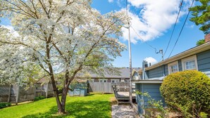 A residential backyard with a stone pathway, a wooden deck, and a tree with white blossoms. - Quaint 3-bedroom cottage in Nashville, shopping, and dining! Pet-friendly! (Nashville)