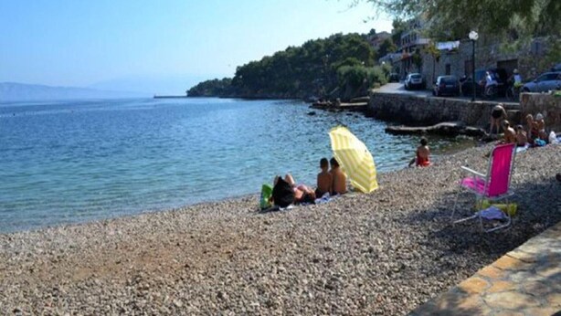 A pebbled beach with people relaxing under umbrellas, a clear view of the sea, and buildings in the background.