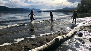 A beach with a wooden log in the foreground, a calm lake, and mountains in the background. - Sunnyside Lakehouse - Semi-waterfront Shuswap Accommodation (Celista)