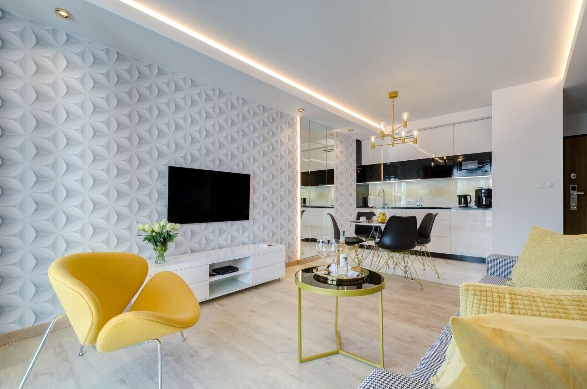 Ein modernes Wohnzimmer mit weißer Wand mit Struktur, einem Flachbildfernseher, gelben Sesseln, einem runden Glastisch und einer Küchenzeile im Hintergrund.