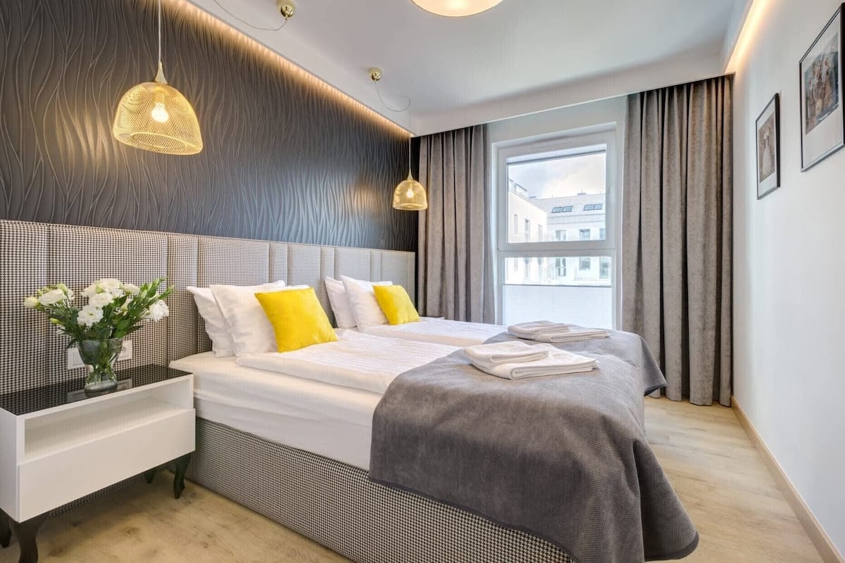 A modern hotel room with a large bed, bedside tables, a vase of flowers, and a view of buildings through the window.