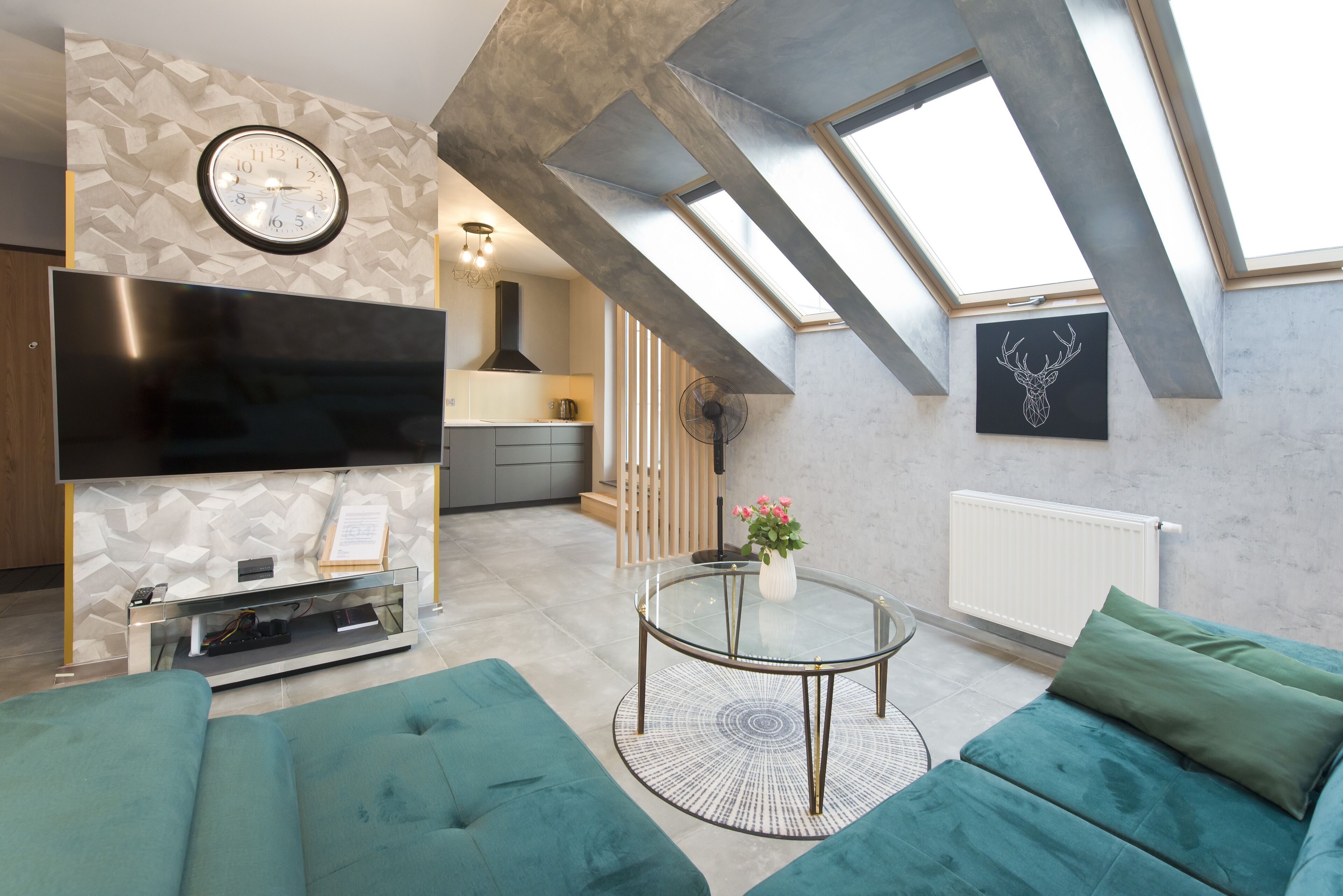 A modern living room with a flat-screen TV, teal sofas, a glass coffee table, and a skylight.