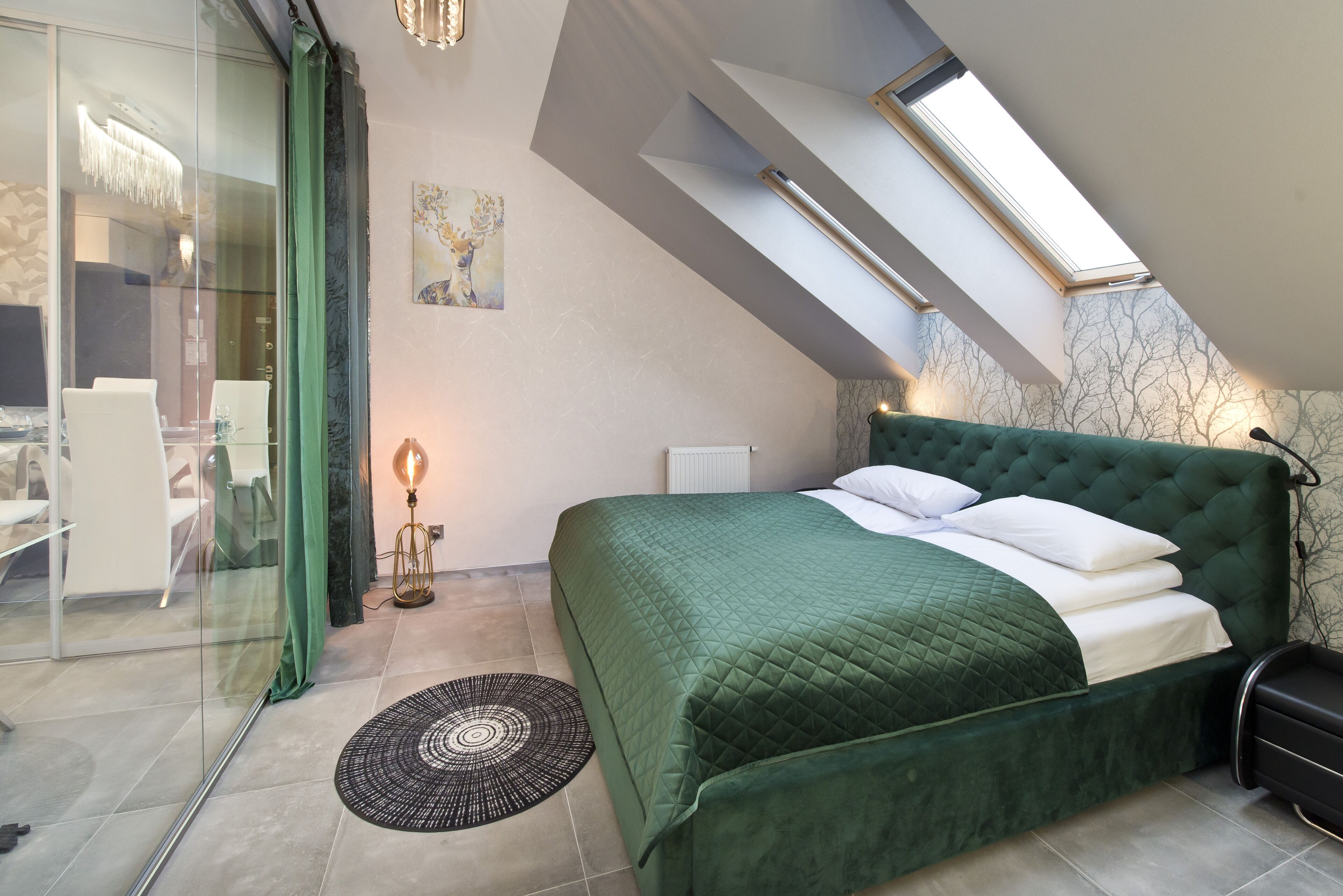 A bedroom with a green bed, a glass wall, a skylight, and a dining area with chairs.