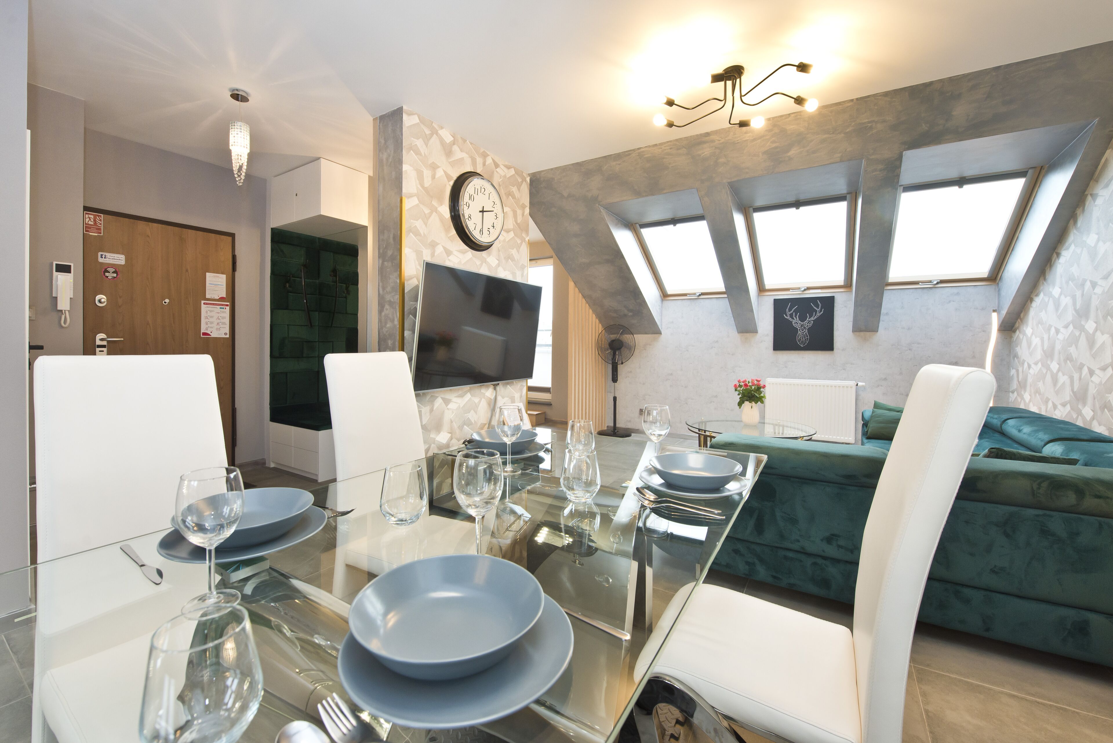 A dining area with a glass table set for four, a clock on the wall, and a green sofa in the background.