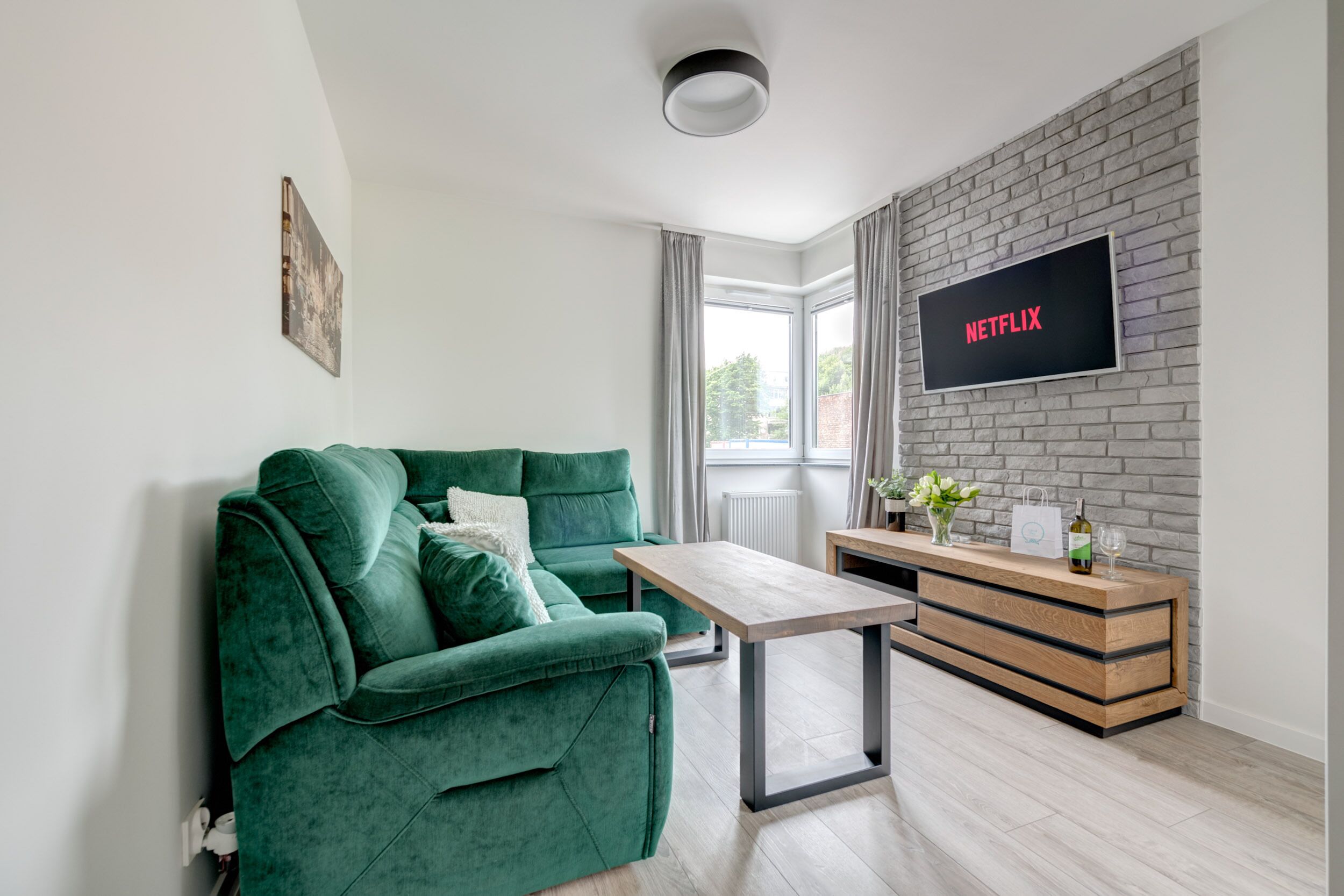 En moderne stue med en grøn sofa, et træbord, et fjernsyn monteret på en murstensvæg og et vindue med gardiner.